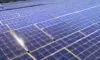 A sweeping panoramic view of a vast solar photovoltaic farm stretching across rolling hills under a golden sunrise, with rows of silver panels reflecting the early light. In the foreground, a maintenance drone hovers above a cluster of panels, its…