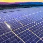 A sweeping panoramic view of a vast solar photovoltaic farm stretching across rolling hills under a golden sunrise, with rows of silver panels reflecting the early light. In the foreground, a maintenance drone hovers above a cluster of panels, its…