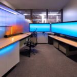 A futuristic home office features a massive custom server rack with glowing blue LEDs against a dark wall. Framed schematics hang nearby, while ambient light illuminates floating dust particles, highlighting the system status display on the front …