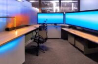 A futuristic home office features a massive custom server rack with glowing blue LEDs against a dark wall. Framed schematics hang nearby, while ambient light illuminates floating dust particles, highlighting the system status display on the front …