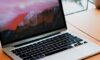 A sleek silver MacBook Pro on a wooden desk bathed in warm morning sunlight, highlighting the glowing Apple logo and metallic texture against a softly blurred office background.