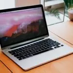 A sleek silver MacBook Pro on a wooden desk bathed in warm morning sunlight, highlighting the glowing Apple logo and metallic texture against a softly blurred office background.