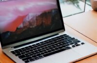 A sleek silver MacBook Pro on a wooden desk bathed in warm morning sunlight, highlighting the glowing Apple logo and metallic texture against a softly blurred office background.