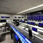 Dimly lit military command center featuring glowing server racks and digital data walls. Operators monitor network topologies under cool blue lighting, illustrating advanced cyber defense infrastructure and technological superiority.