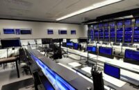 Dimly lit military command center featuring glowing server racks and digital data walls. Operators monitor network topologies under cool blue lighting, illustrating advanced cyber defense infrastructure and technological superiority.