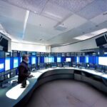 Futuristic security command center with analysts monitoring large curved screens displaying real-time certificate trust metrics and automated renewal dashboards under soft blue lighting, highlighting human oversight in PKI management.