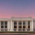 High‑resolution photorealistic view of the Pentagon’s white marble façade bathed in a dramatic sunset, with faint holographic AI circuitry patterns glowing across the building, illustrating the convergence of defense and technology under cinematic…