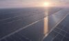 Sunrise over Gujarat data center campus with silver server halls, expansive solar farms, wind turbines, green desert vegetation, and high‑voltage transmission lines