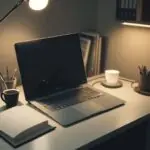 A cozy home office desk featuring a laptop displaying a terminal window with code, a notebook with a pen, and a warm cup of coffee under a soft desk lamp.